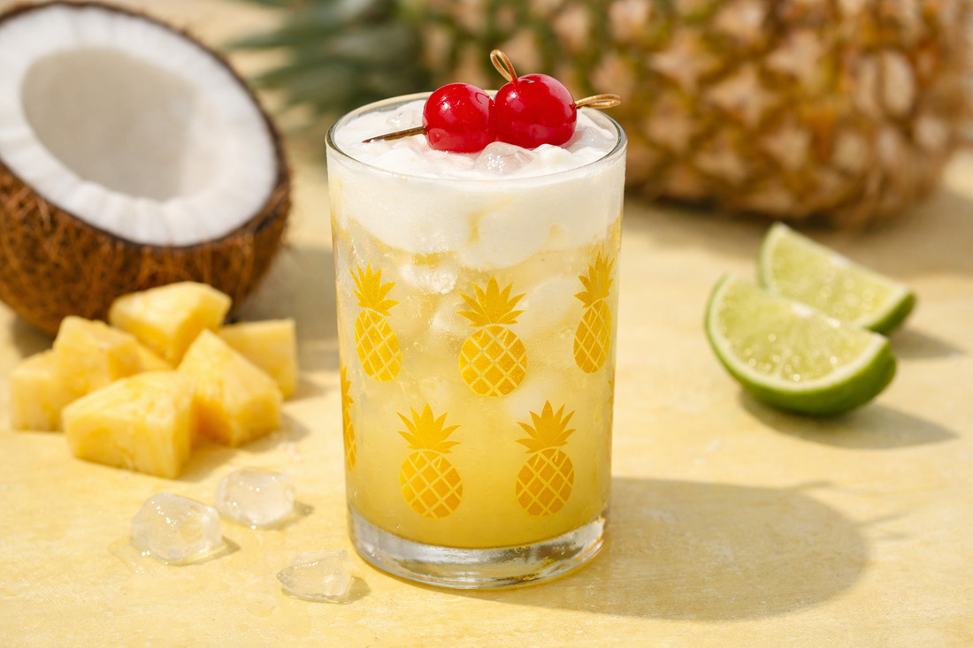 A tropical Dirty Kava Colada mocktail with a pale yellow base and creamy float, topped with maraschino cherries in a pineapple-patterned glass, set against a light yellow background with fresh pineapple, coconut, and lime.
