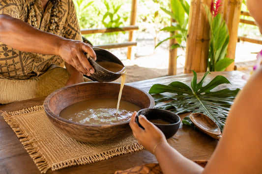 Traditional kava preparation with kava being poured from a coconut shell into a serving cup beside a wooden bowl in a South Pacific-inspired setting.