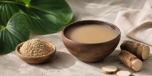 Minimalist still-life image of a traditional kava root drink in a rustic wooden bowl with creamy, earthy-toned liquid, surrounded by kava root pieces and ground kava powder on a neutral textured surface with soft tropical greenery in the background.