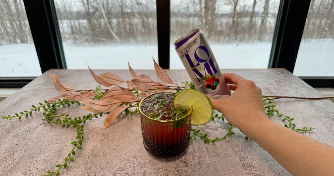  Glass of tart cherry and mint mocktail garnished with lime, served beside a can of Lomi Berry Breeze by a window with winter scenery.