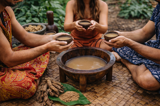 Group participating in a traditional kava ceremony, seated on woven mats and holding coconut shell cups around a communal kava bowl in a natural outdoor setting.